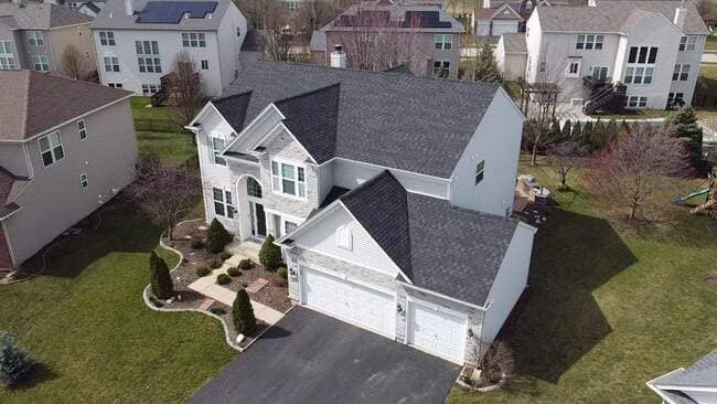 Elevated diagonal view of a large, modern two-story suburban home featuring a newly installed dark gray and black architectural shingle roof. The house has light siding and stone veneer accents on the facade and attached three-car garage. The complex, multi-gabled roof system showcases a comprehensive premium roof upgrade by professional roofing specialists under a clear sky.